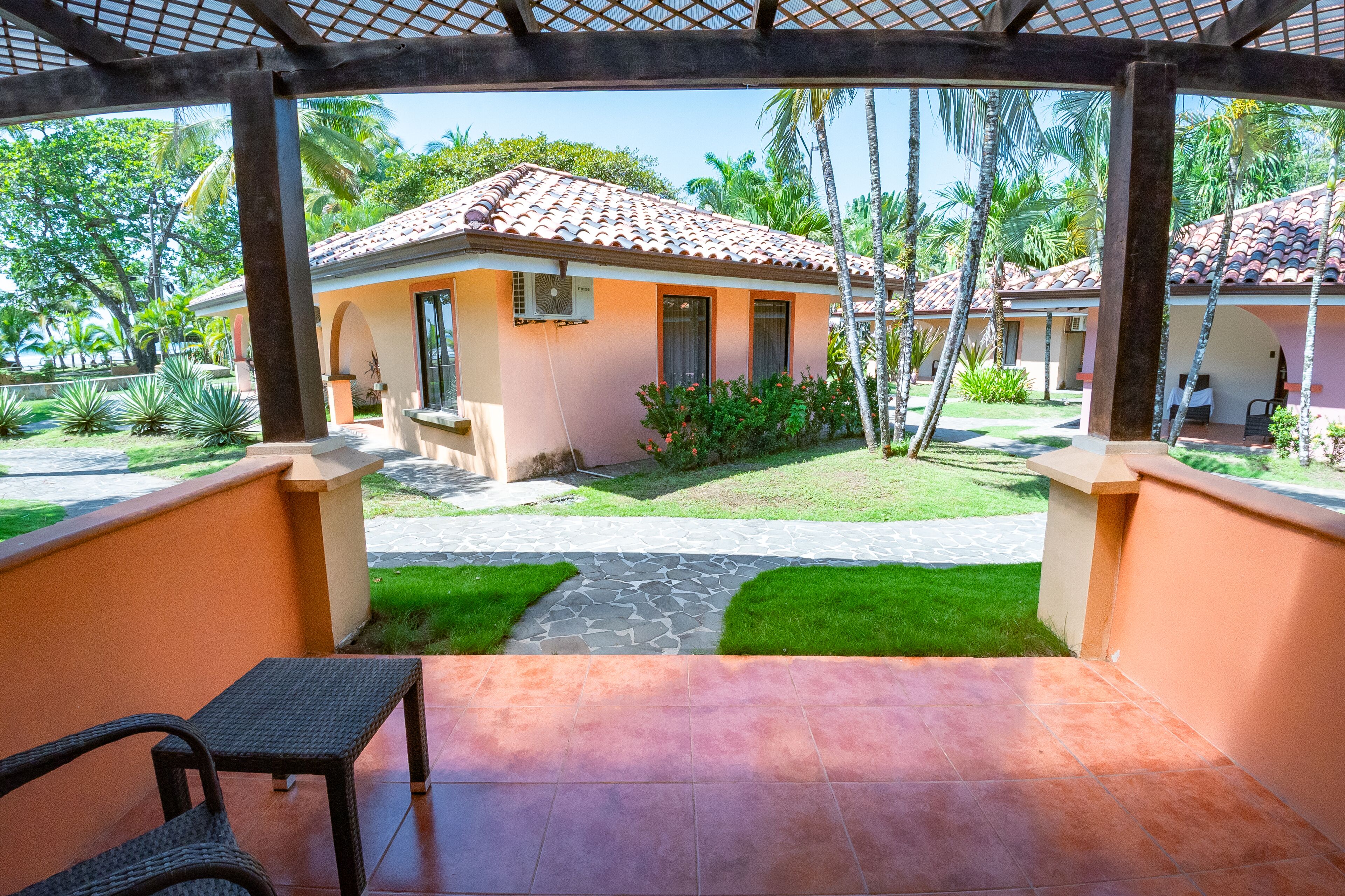 Un patio cubierto con vistas a un edificio de techo de tejas, rodeado de vegetación y palmeras.
