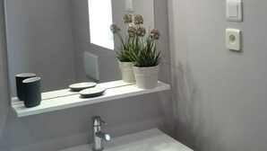 Une salle de bain avec un lavabo blanc, un miroir, une plante en pot et un interrupteur sur le mur.