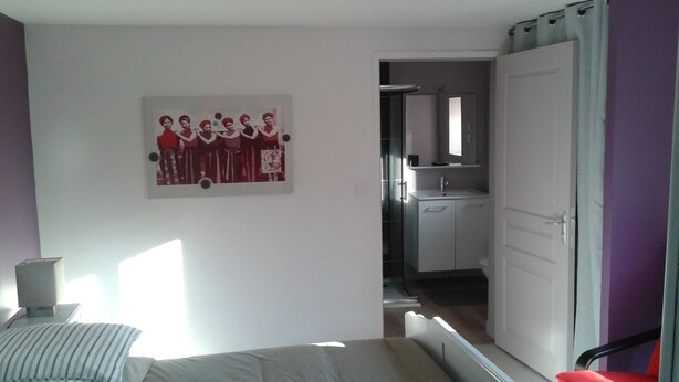 Une chambre à coucher avec un lit, une chaise, un tableau, et une vue sur la salle de bain et la cuisine.