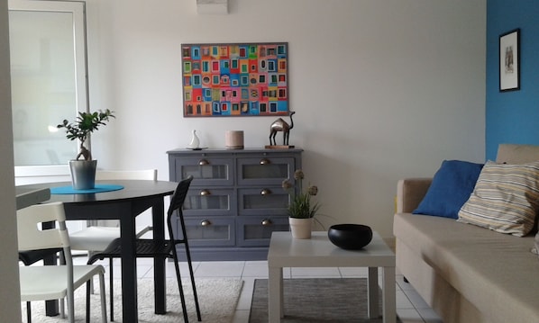 Un salon avec un mur bleu, un canapé, une table basse, une table à manger avec des chaises, un meuble et un tableau abstrait aux couleurs vives.