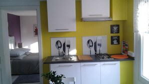 Une cuisine de taille réduite avec des meubles blancs, une cuisinière à gaz et un évier. Des tableaux encadrés représentant des couverts sont accrochés au mur.