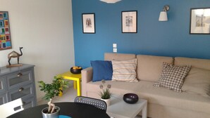 Un salon avec un mur bleu, un canapé beige, une petite table et une chaise. Des tableaux encadrés ornent le mur et une petite plante est posée sur la table.
