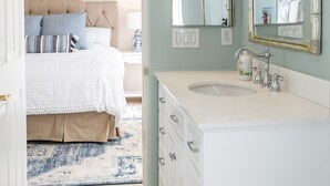 A bedroom with a bed, bedside table, and a bathroom with a sink, mirror, and cabinets. - Desert Oasis on PGA WEST Palmer Private - #100182 3BR (La Quinta)