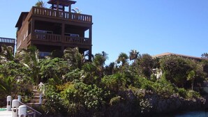A multi-story building with balconies, a wooden deck, and a rocky shoreline with palm trees. - Waterfront Luxury 3 Bedroom w/ Pool and Dock (West End)