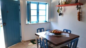 A dining area with a wooden table, blue chairs, a window with blue shutters, and a wall-mounted shelf with kitchen utensils.