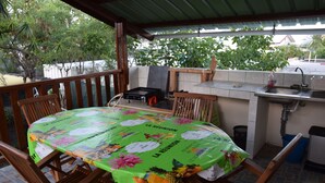 Un espace extérieur abrité, aménagé pour prendre les repas, avec une table recouverte d’une nappe, des chaises en bois, un petit coin cuisine avec un évier et une poubelle.