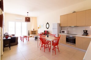 A modern kitchen with wooden cabinets, a microwave, and a coffee maker. There is a dining area with red chairs and a table.