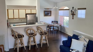 A modern kitchen with a central island, bar stools, a dining area with a table and chairs, and a living room with a blue sofa.