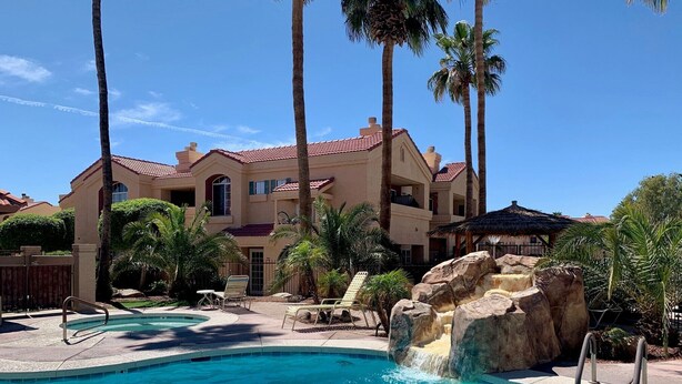 A pool with a waterfall feature, surrounded by palm trees and a large building with a red-tiled roof.
