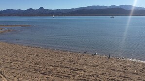 A beach with a clear view of mountains in the background, a calm lake, and a few boats.
