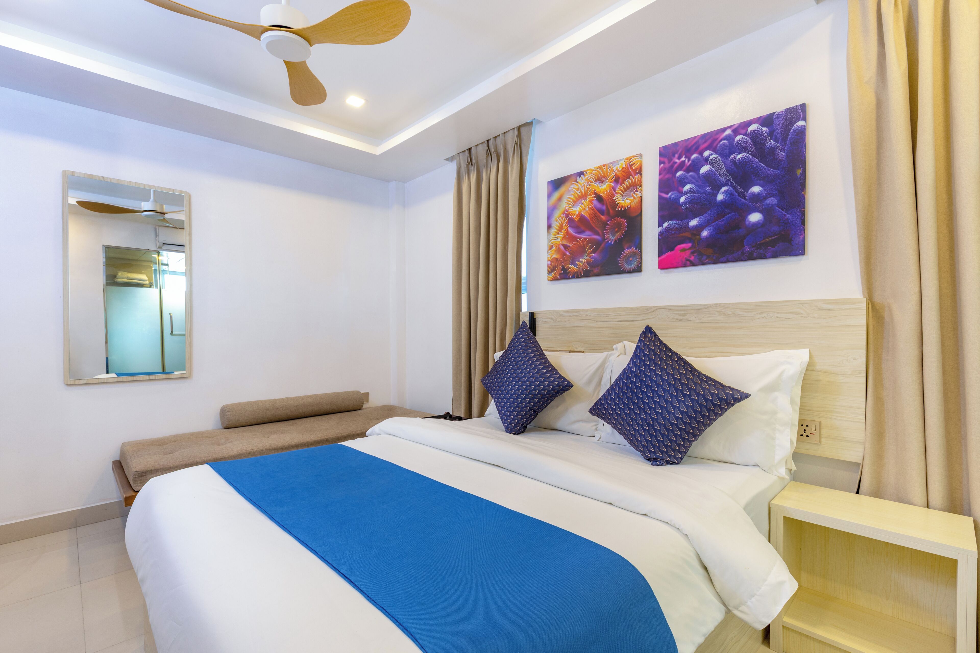 A modern hotel room with a large bed, a ceiling fan, a bedside table, a mirror, and two wall art pieces.
