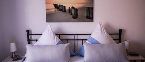 Une chambre d’hôtel avec un lit, des oreillers, une table de chevet, une lampe et un tableau représentant un coucher de soleil sur une plage avec des poteaux en bois.