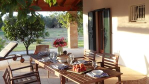 Um pátio externo com mesa de madeira posta para refeições, cadeiras de vime e vista para o jardim.