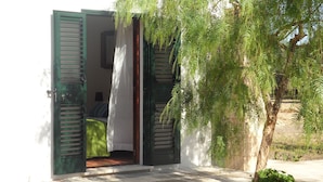 Um quarto com uma cama, uma janela com persianas verdes e uma árvore lá fora.