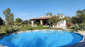 Piscina com área de terraço, cercada por vegetação e um pequeno edifício com telhado telhado.