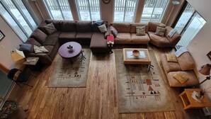 Un salon spacieux avec un grand canapé marron en forme de L, deux tables d’appoint et une table basse. La pièce est dotée d’un parquet et de grandes fenêtres qui laissent entrer la lumière naturelle.