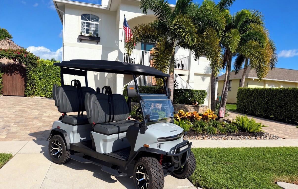 A golf cart parked in front of a residential building with a flag, palm trees, and a well-maintained lawn.