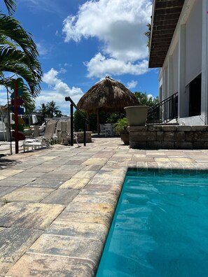 Uma área de piscina com estrutura de telhado de palha, cadeiras de lounge e um caminho de pedra que leva à piscina.
