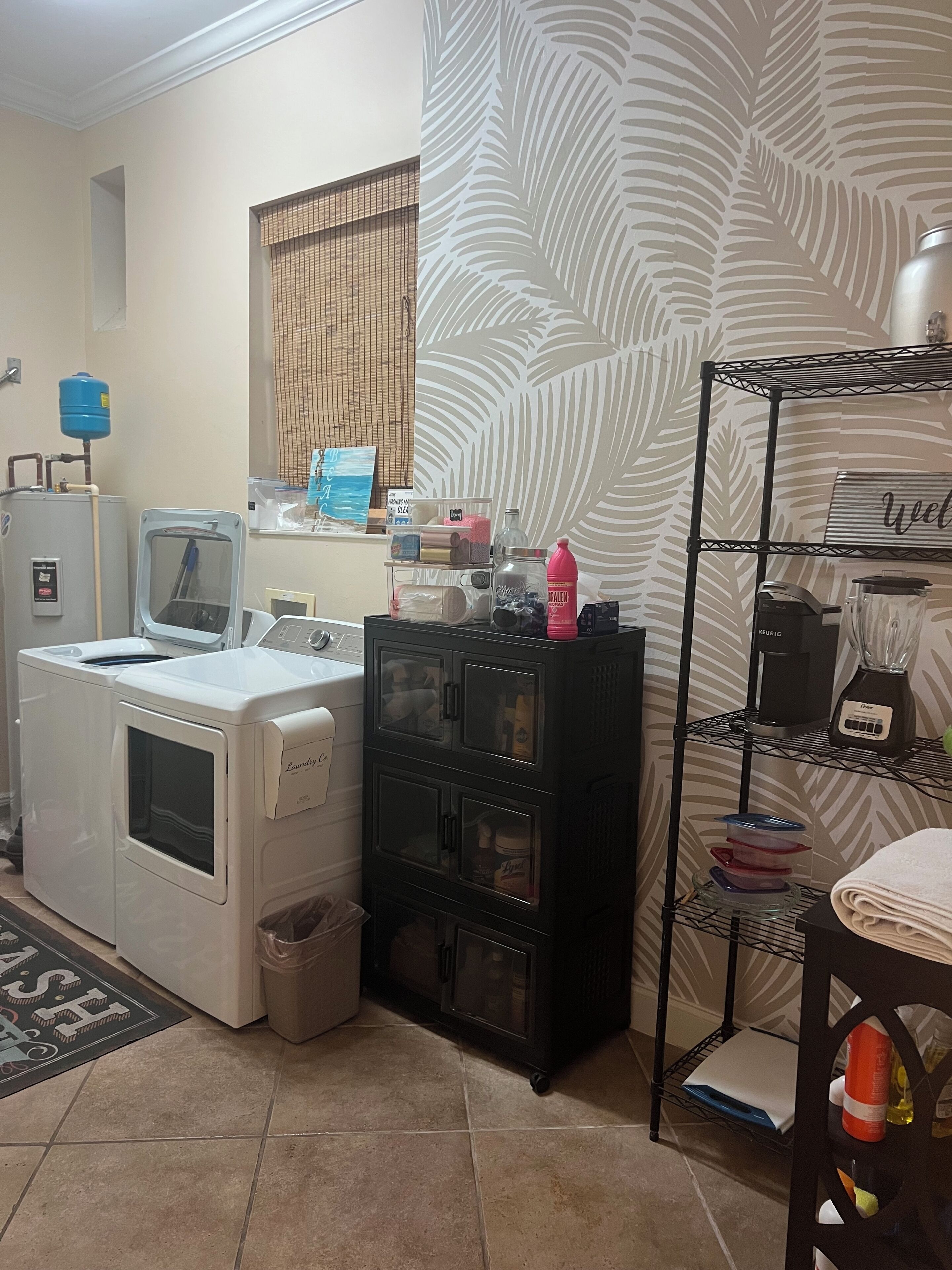 A laundry area with a washing machine, dryer, and a water heater. There is a black storage unit with shelves and a metal rack with a coffee maker and a blender.