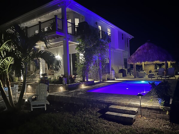 Uma casa de dois andares com piscina, área de estar externa e uma construção com telhado de palha.