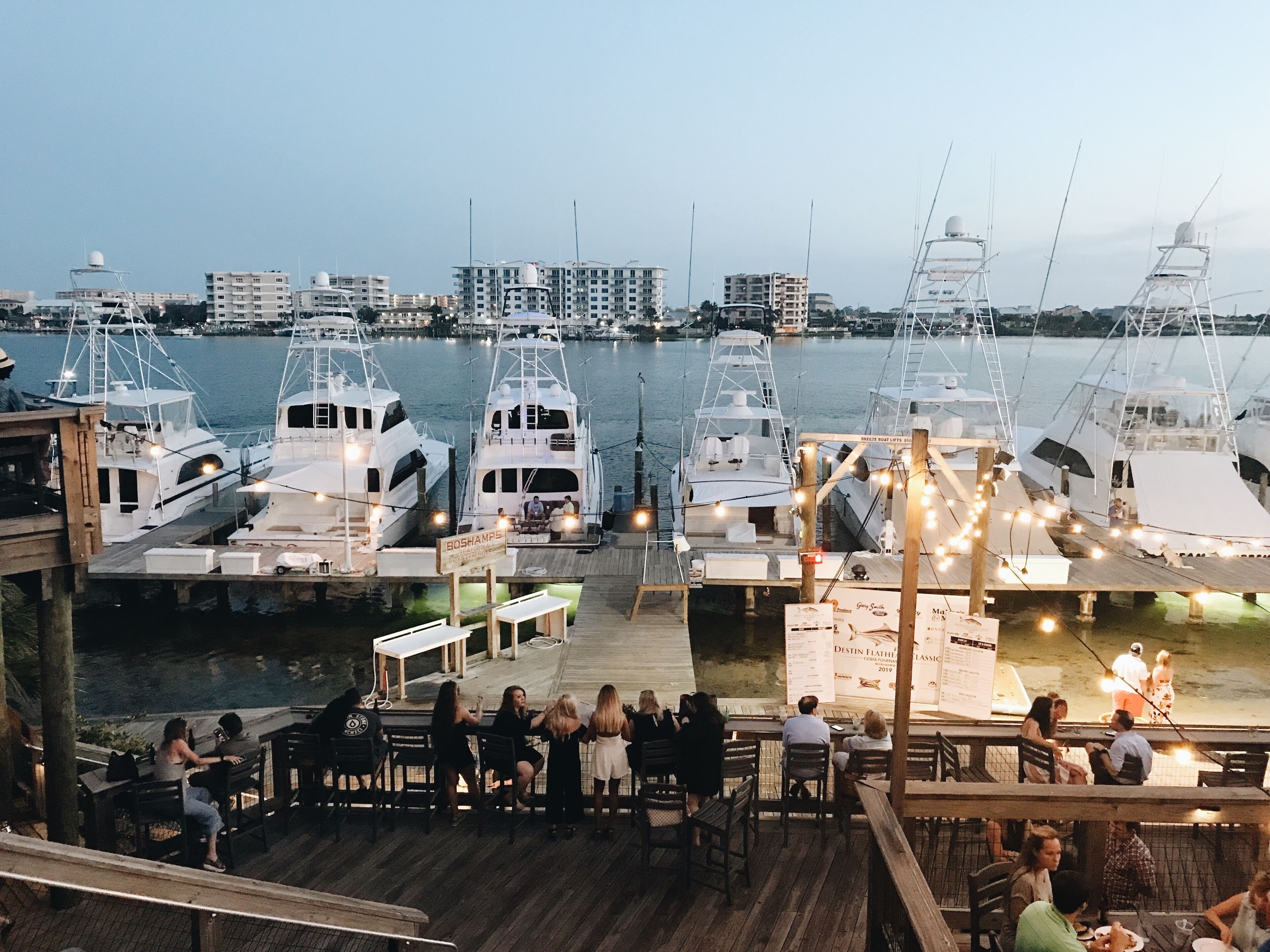 A marina with multiple yachts docked, a wooden deck with tables and chairs, and a view of the water.