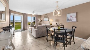 A spacious living area with a dining table, chairs, and a sofa. There is a view of the outdoors through large windows. -  Perfect location! Ground floor, 40 Steps to Pool or Beach! Pets Welcome! (Miramar Beach)
