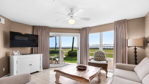 A living room with a television, a wooden coffee table, two armchairs, a sofa, a ceiling fan, and a view of a body of water through large windows. -  Perfect location! Ground floor, 40 Steps to Pool or Beach! Pets Welcome! (Miramar Beach)