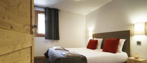 A hotel room with a wooden door, a bed with white and red pillows, a bedside table with a lamp, and a window with curtains.