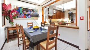 Un coin repas avec une nappe à carreaux, des chaises en bois, un panier de fruits et un tableau coloré accroché au mur.