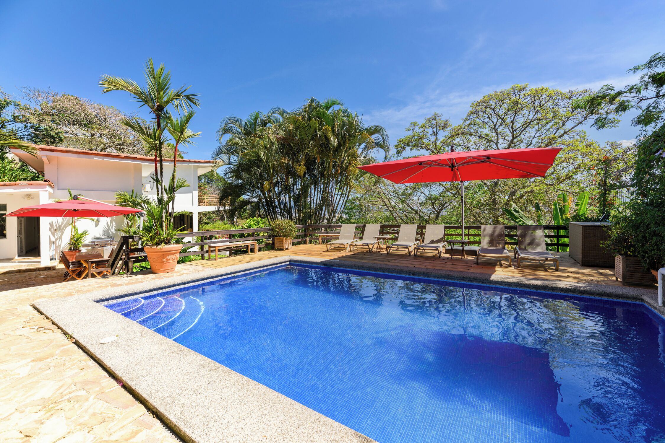 A swimming pool with a tiled edge, surrounded by lounge chairs and umbrellas, with a wooden deck and potted plants.