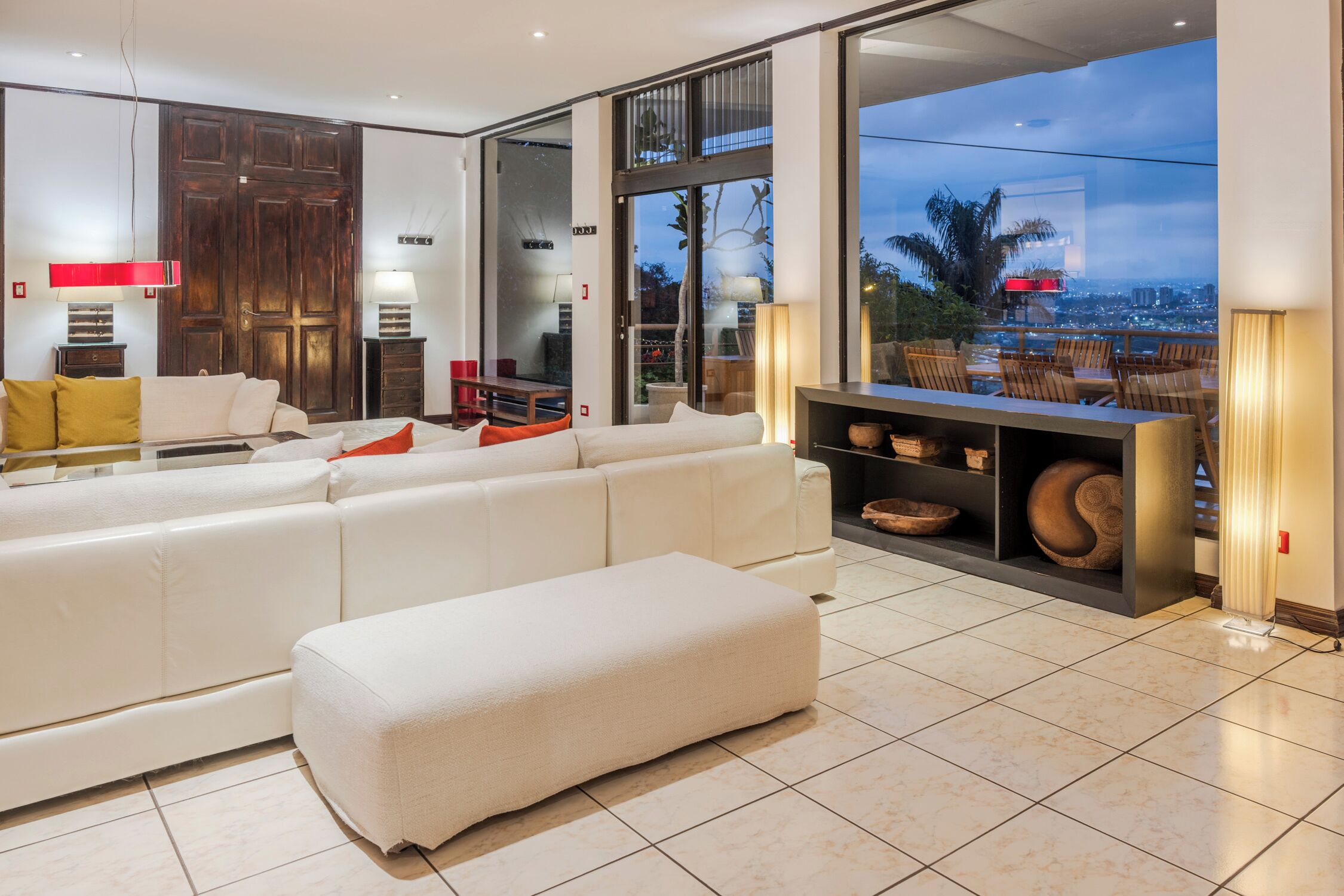 A modern living room with a large white sofa, a small ottoman, and a glass door leading to a balcony with a city view.