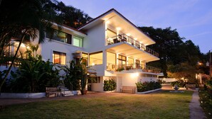 Une maison moderne à deux étages, dotée d’un balcon, entourée d’une végétation luxuriante et d’une pelouse bien entretenue.