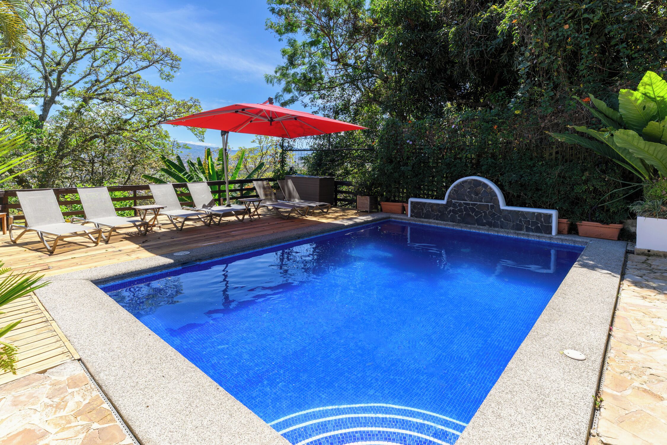 Una piscina con borde de azulejos, rodeada de sillas de descanso y un paraguas rojo.