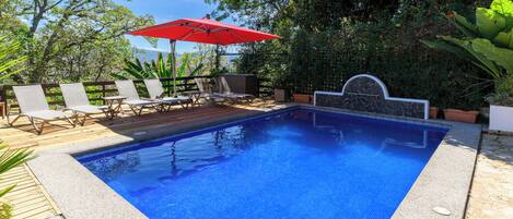 Una piscina con borde de azulejos, rodeada de sillas de descanso y un paraguas rojo.