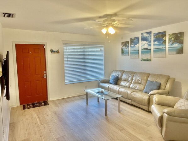 A living room with a beige leather sofa, a glass coffee table, and a ceiling fan. There are three framed pictures on the wall.