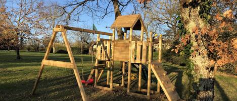 Una estructura de juego de madera con columpios y un tobogán, rodeada de árboles y césped.