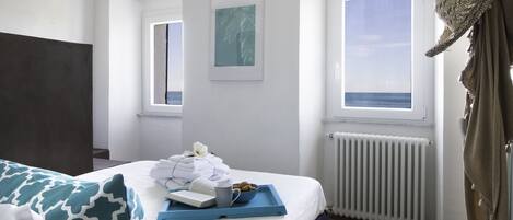 A hotel room with a bed, a tray with towels and a book, a framed picture on the wall, and a view of the sea through a window.