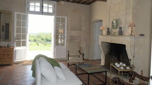 Una habitación con un sofá blanco, una mesa con libros, una chimenea y vistas al exterior.