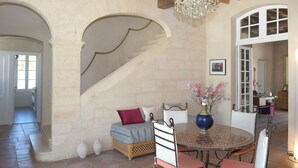Una habitación amplia con pared de piedra, una escalera, un candelabro, una mesa redonda y una cama con cubrecama estampado.