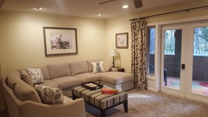 A living room with a beige sectional sofa, a small striped ottoman, a framed picture on the wall, a lamp, and a sliding glass door leading to a patio.