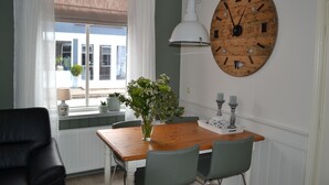 A dining area with a wooden table, two chairs, a vase with flowers, a wall clock, and a window with curtains. - Apartment in the middle of the center of Oostkapelle an apartment with charm (Oostkapelle)