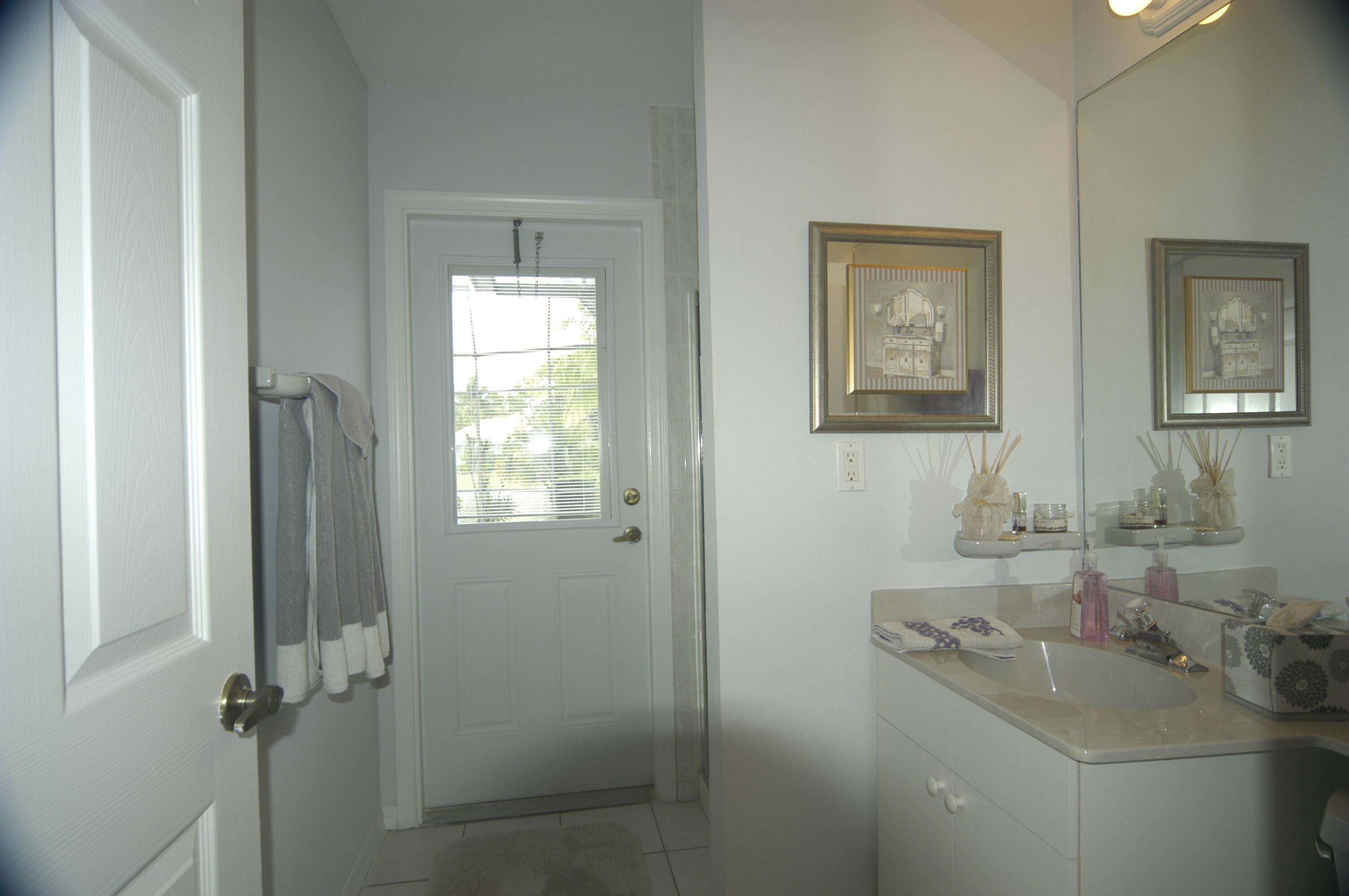 A bathroom with a white door, a window with blinds, a sink with a mirror, and a shelf with toiletries.
