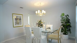 A dining area with a glass table, white chairs, a chandelier, a framed artwork, and a potted plant.
