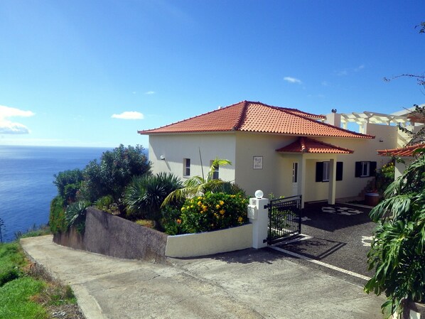 A coastal house with a red-tiled roof, a paved driveway, and a garden with yellow flowers.