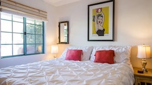 Habitación de hotel con una cama grande, dos almohadas rojas, una obra de arte enmarcada sobre la cama, una ventana con persianas y una mesita de noche con una lámpara.