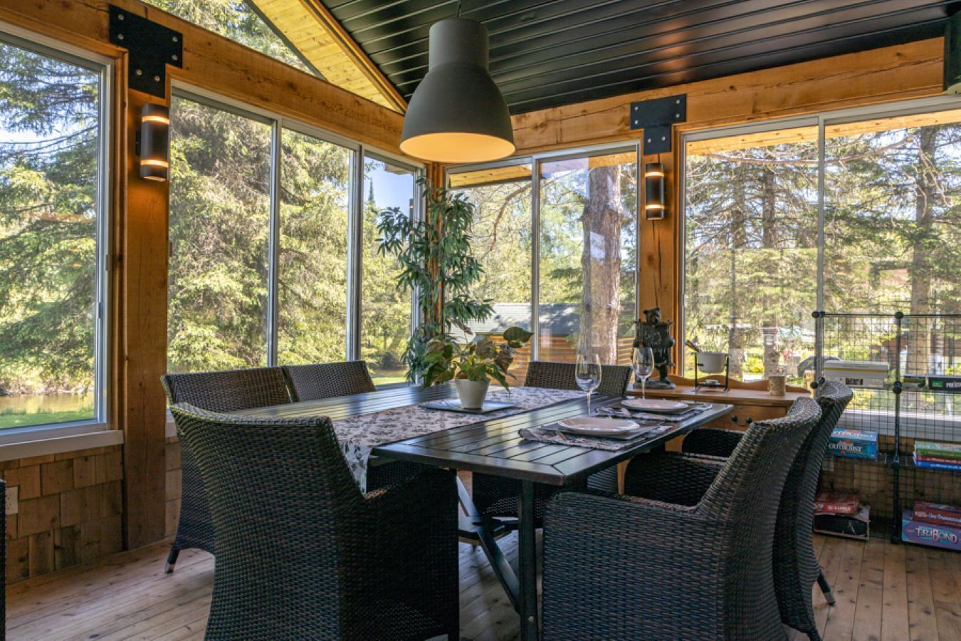 A dining area with a table set for four, surrounded by wicker chairs, large windows offering a view of trees, and a hanging pendant light.