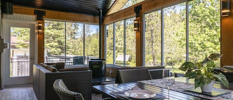 Eine überdachte Terrasse mit Korbmöbeln, einem Esstisch für zwei Personen und großen Fenstern mit Blick auf die umliegenden Bäume.