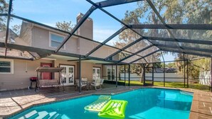 Une piscine dans le jardin, avec un patio couvert, une chaise suspendue et une petite table.