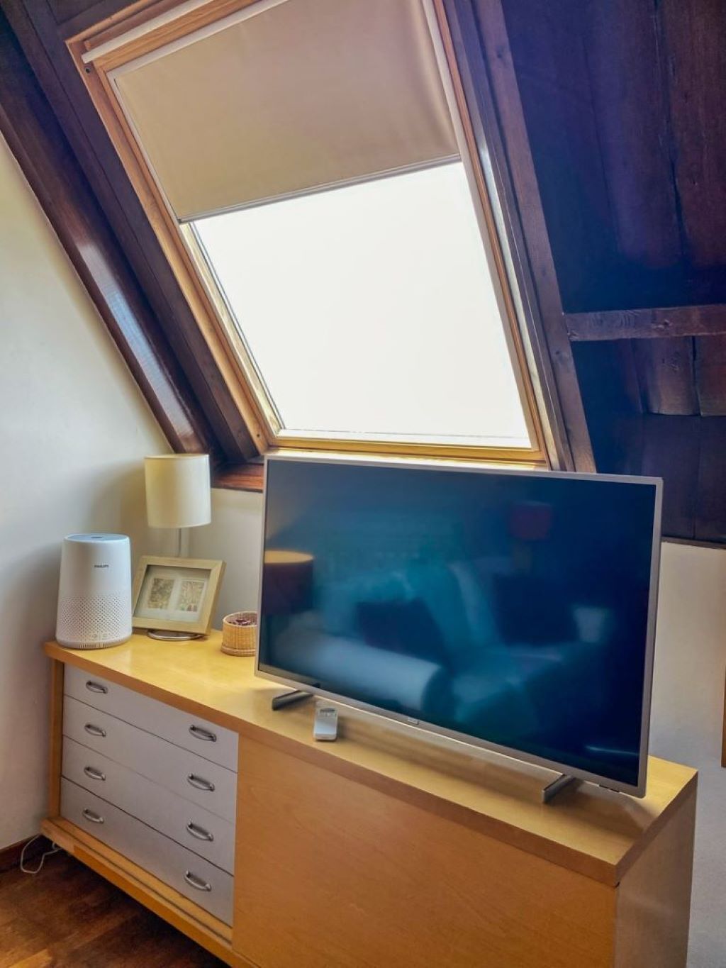 Un téléviseur posé sur un meuble en bois, avec un cadre photo et une lampe à côté, le tout sous une lucarne.