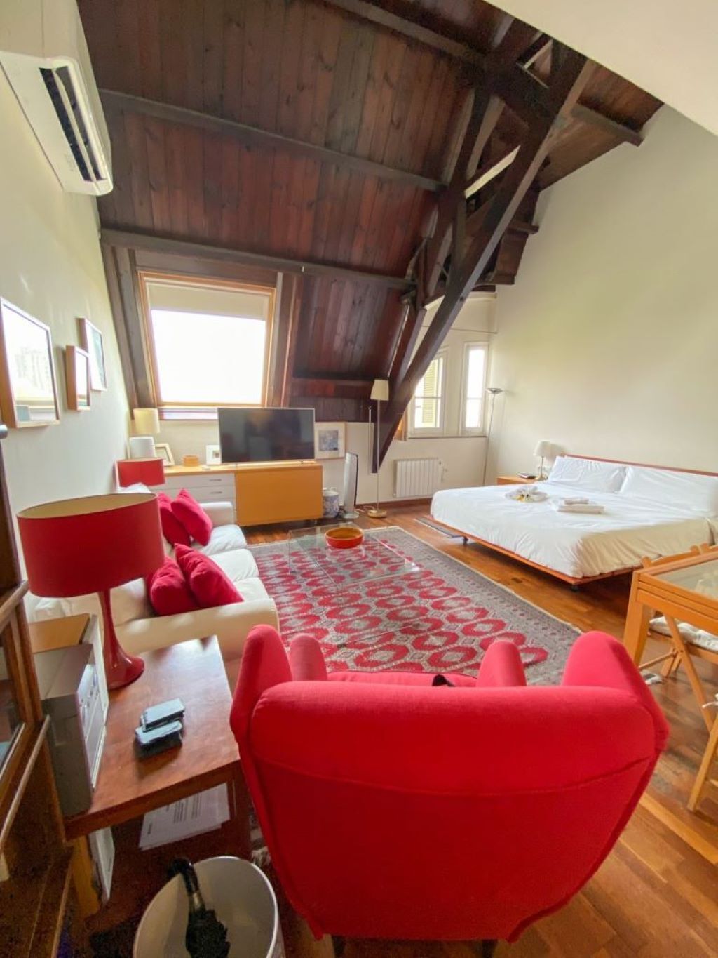 Une pièce confortable avec un plafond en bois, un lit, une télévision, un fauteuil rouge et un bureau avec une lampe.